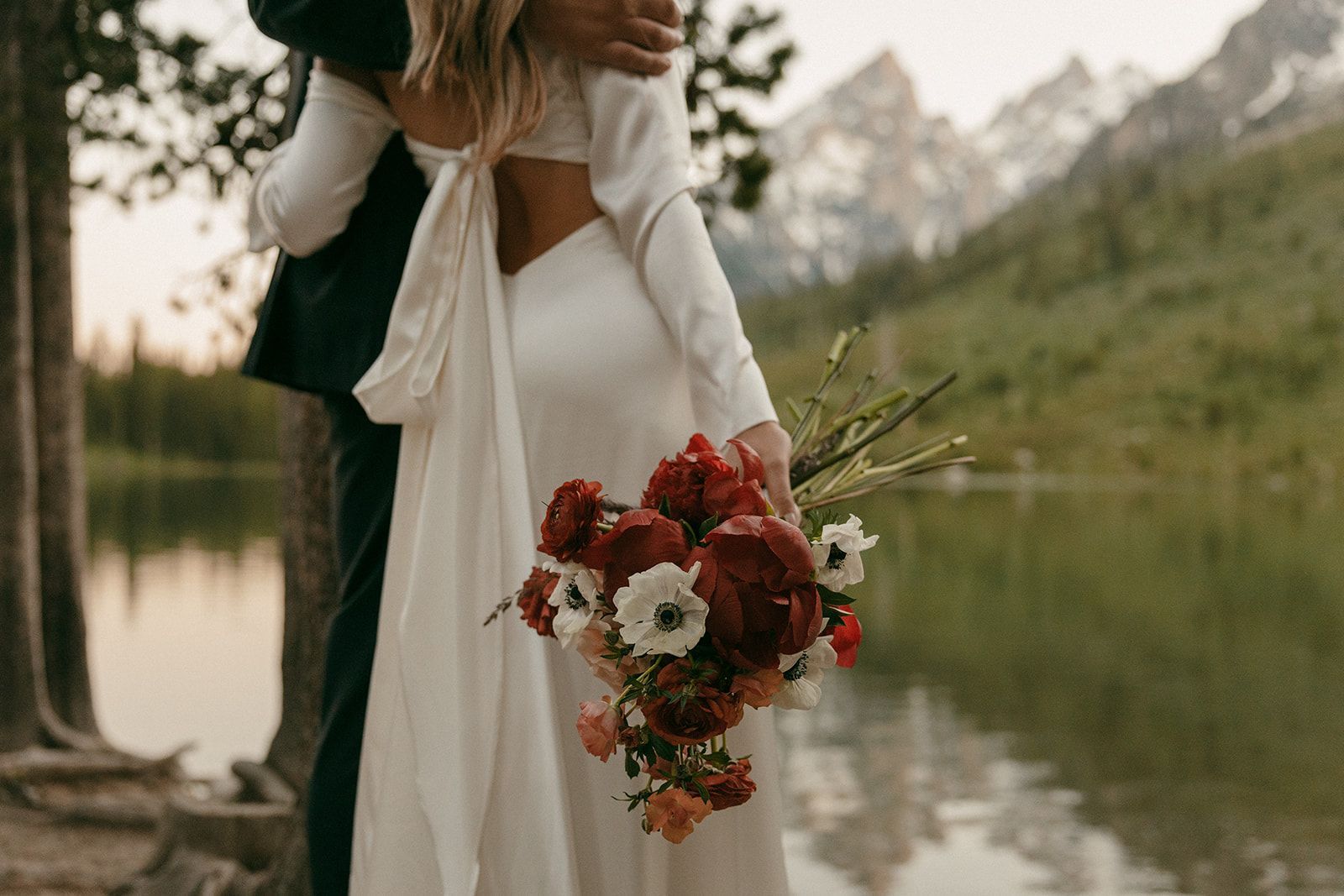Bride in open-back dress with bouquet, embraced by groom, lake and mountains in background.