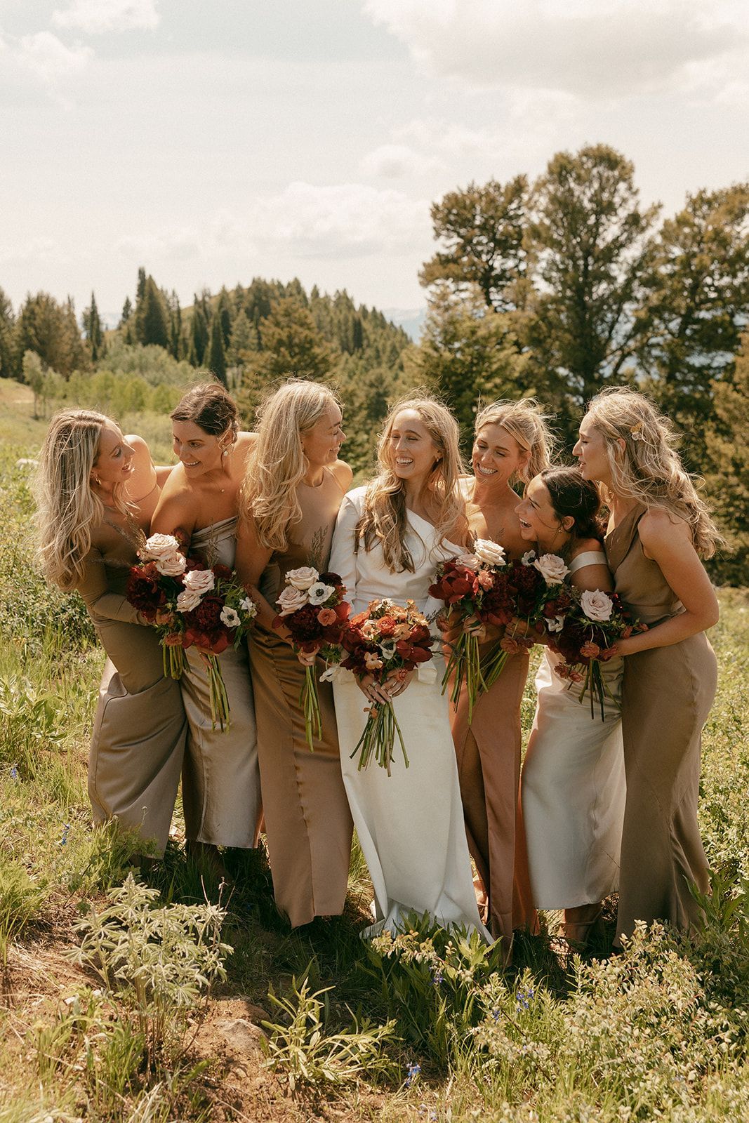 Bride and bridesmaids in dresses, holding bouquets, laughing in a field. Brown, gold, and white. Trees in background.