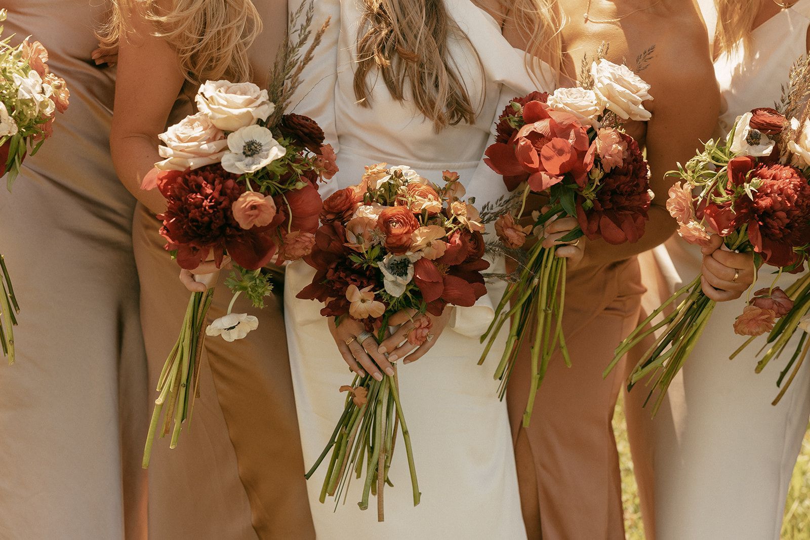 Bridesmaids in tan and white dresses holding bouquets of red and cream flowers.