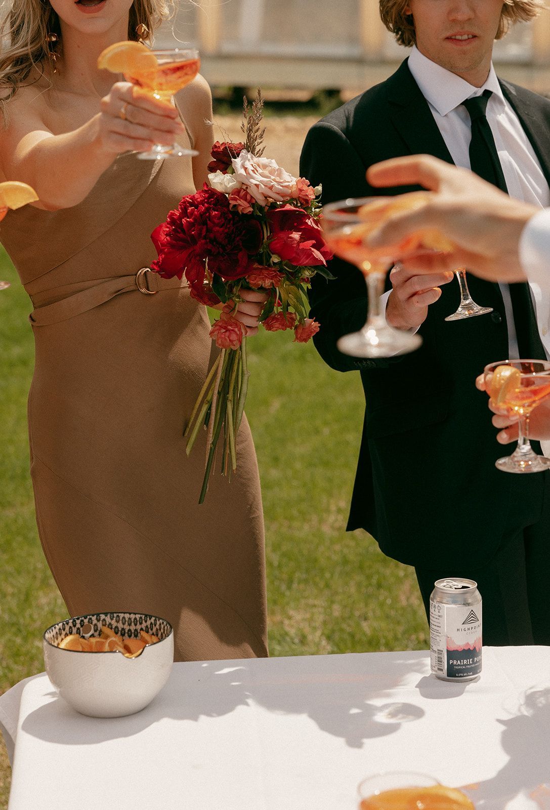People toasting drinks outdoors, woman holding flowers, man in suit.