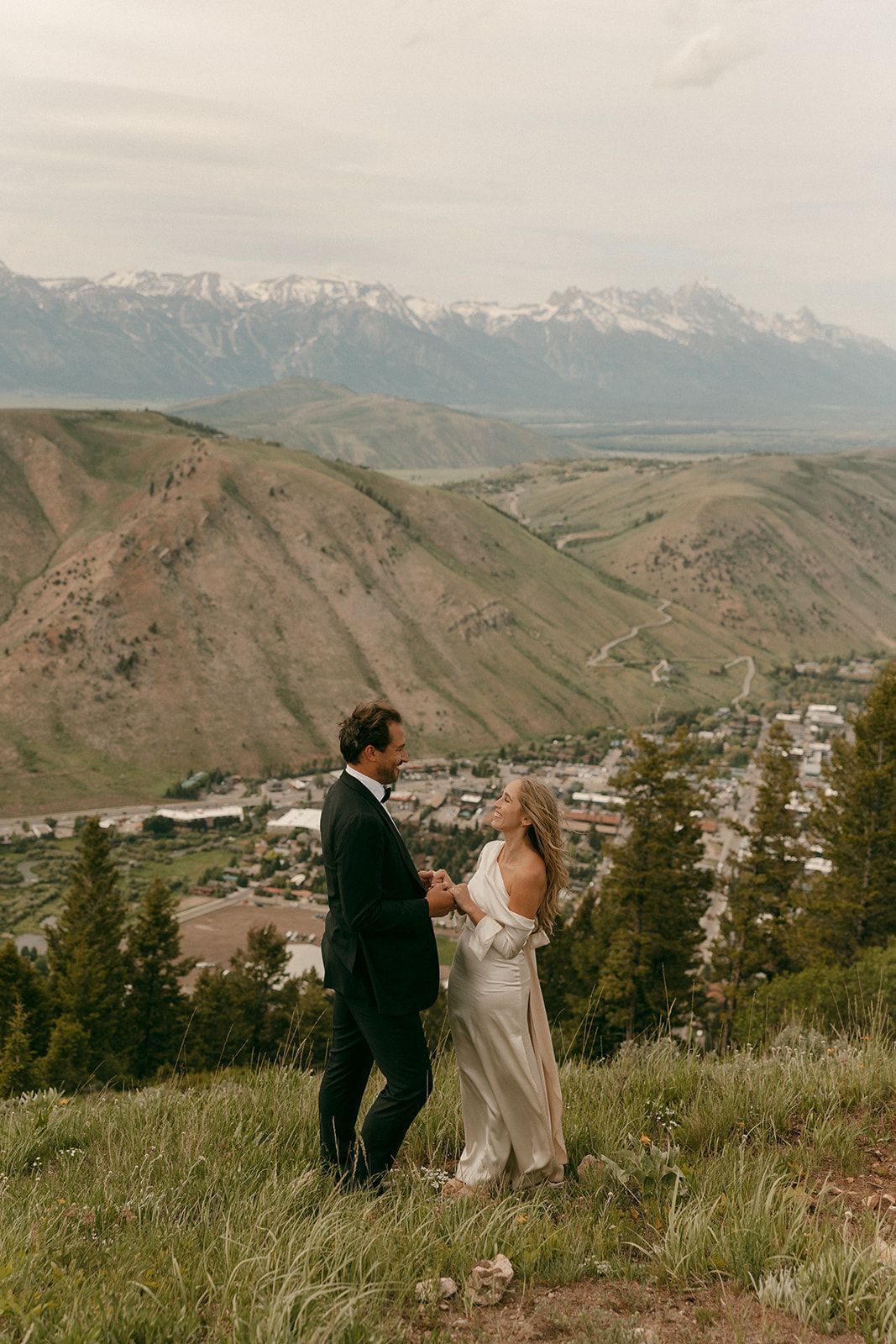 Couple holding hands on a hillside overlooking a town, mountains in background.