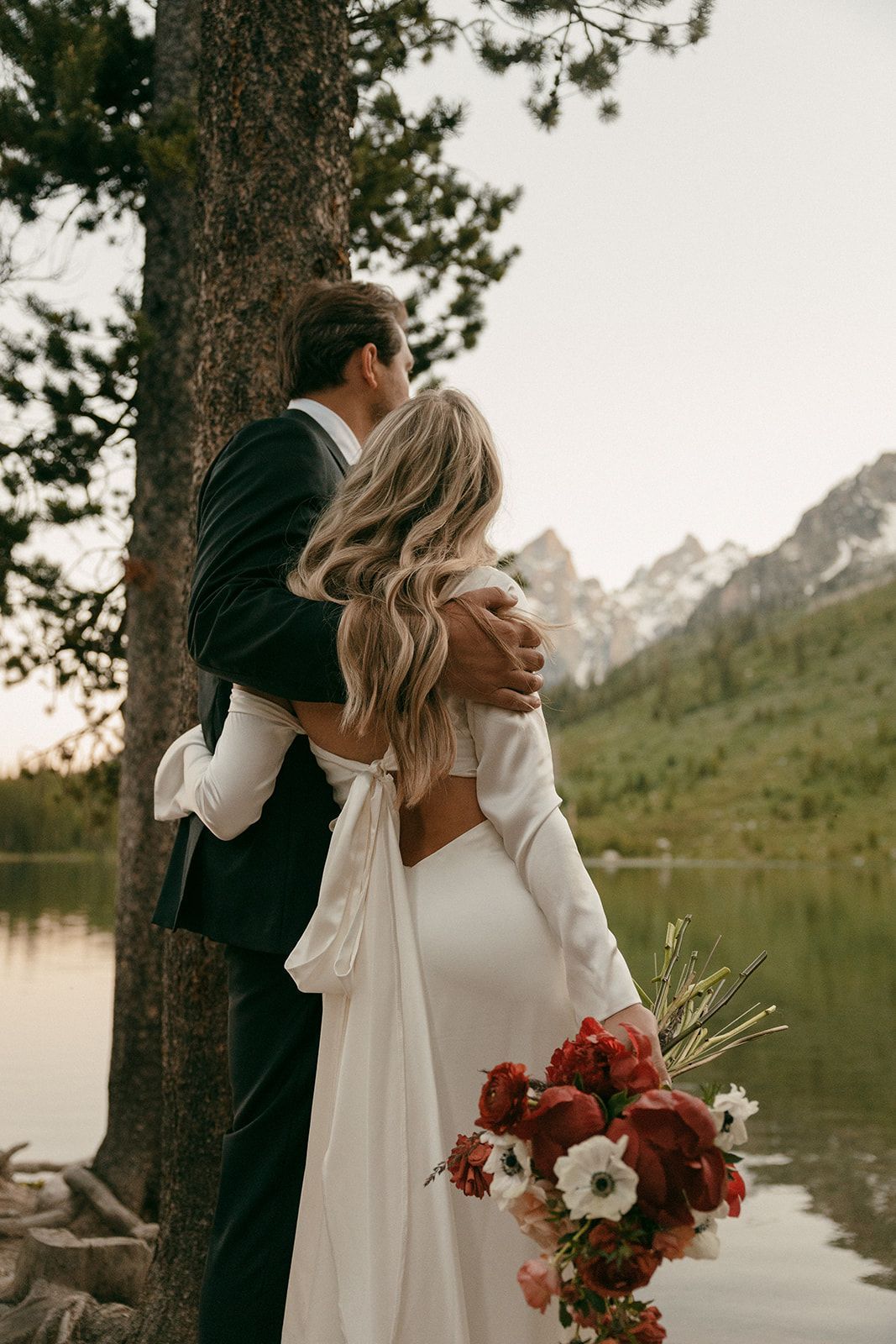 Couple embracing near lake, with mountain backdrop. Woman in white dress holds bouquet; man in suit.
