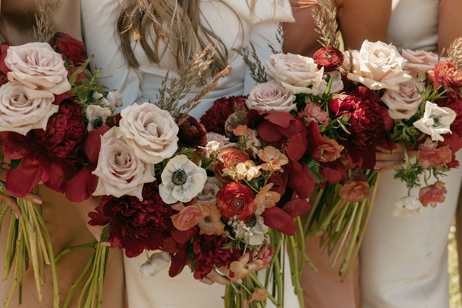 Bridesmaids holding bouquets of blush roses and burgundy flowers with wheat, outside.