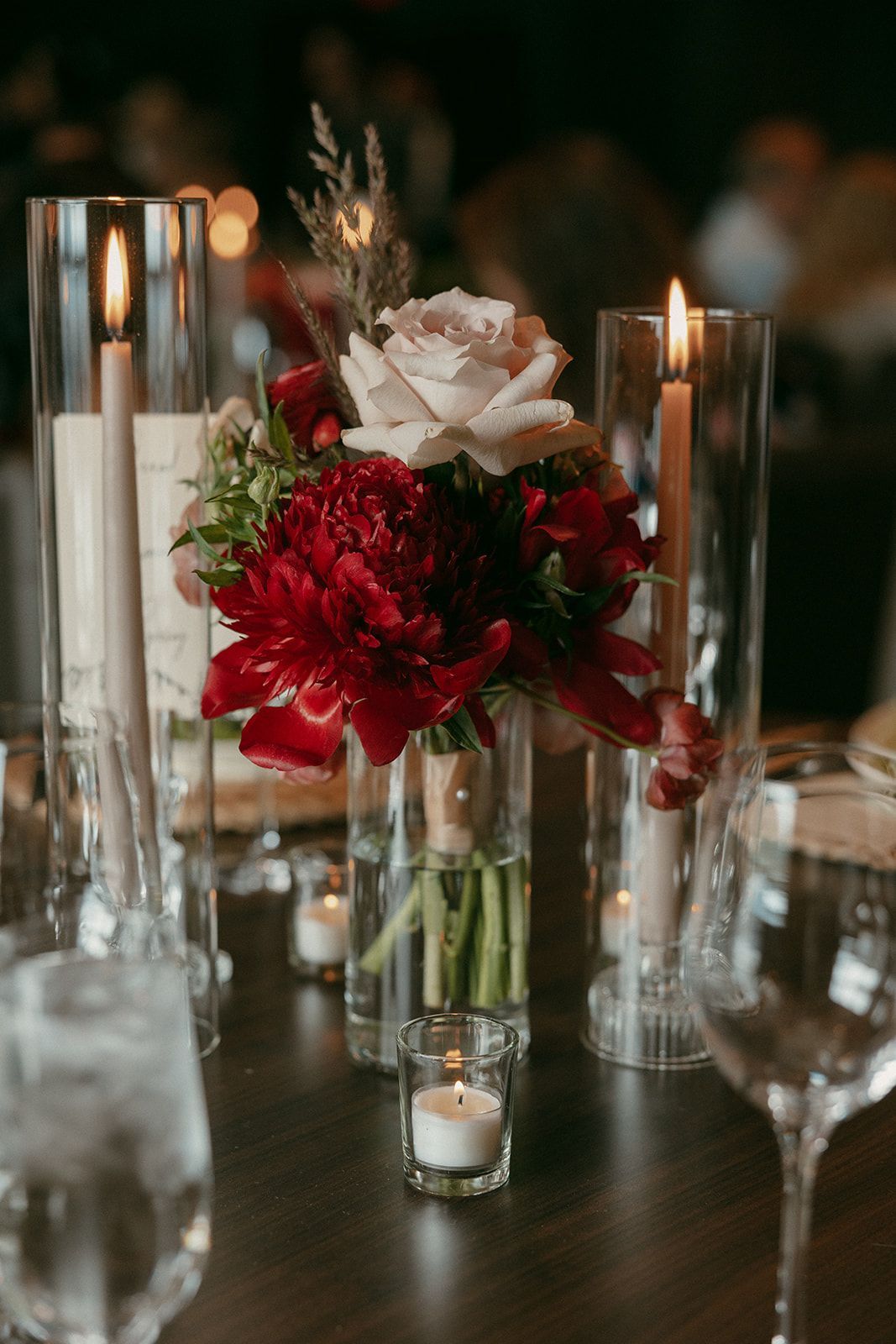 Centerpiece with red flowers, white rose, candles in glass vases, and glassware on a dark wooden table.