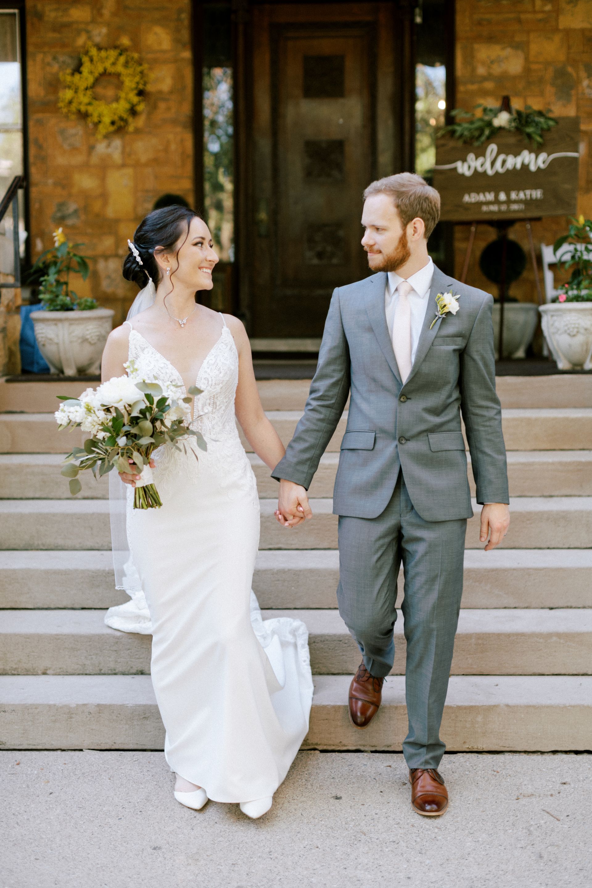 Bride and groom holding hands, exiting a building with stone steps. Bride in white gown, groom in gray suit, smiling.