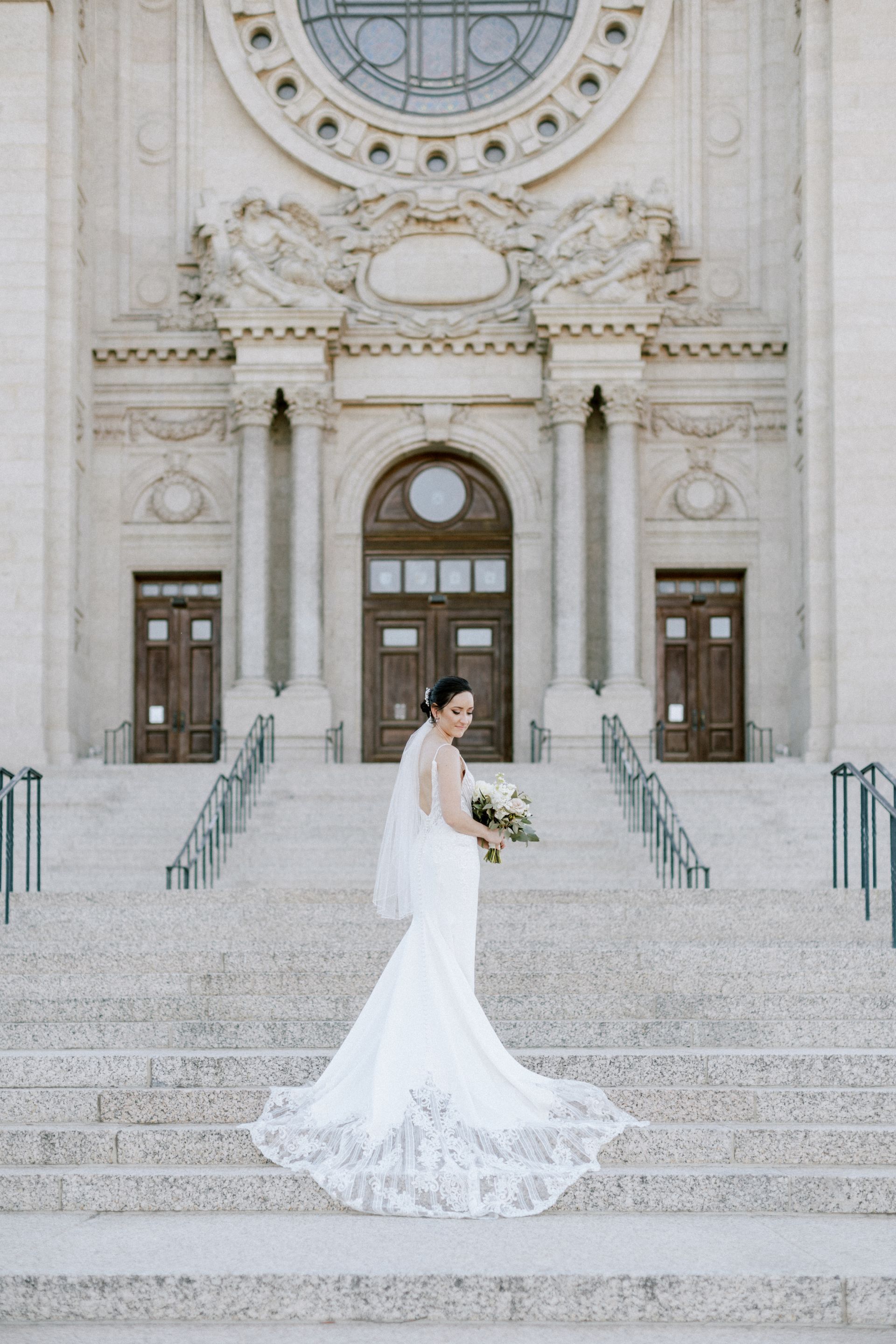 Bride in a white gown stands on steps in front of a large stone building, holding a bouquet, looking back.