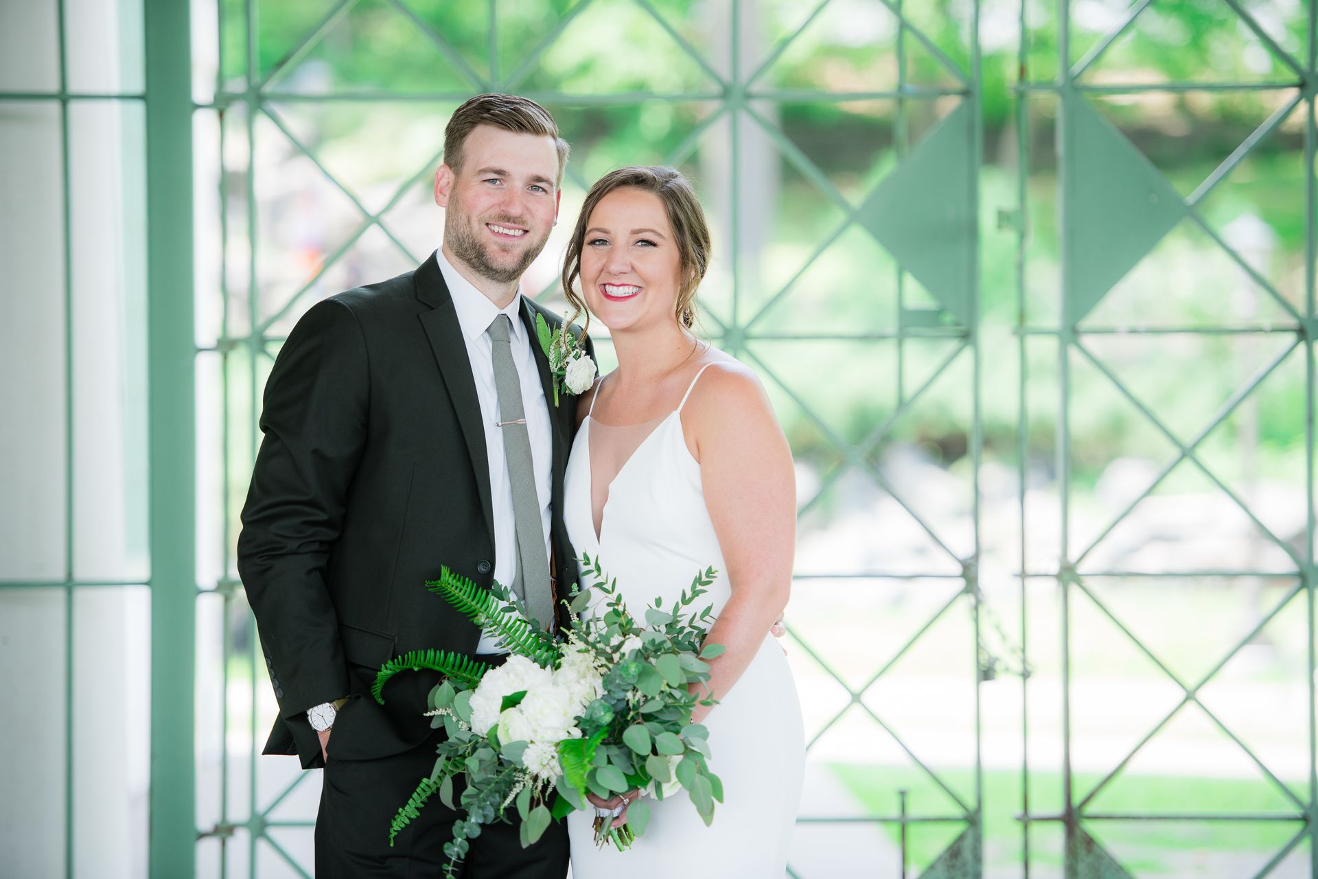 Couple in wedding attire smiling, posing in front of decorative window. The bride holds a bouquet.