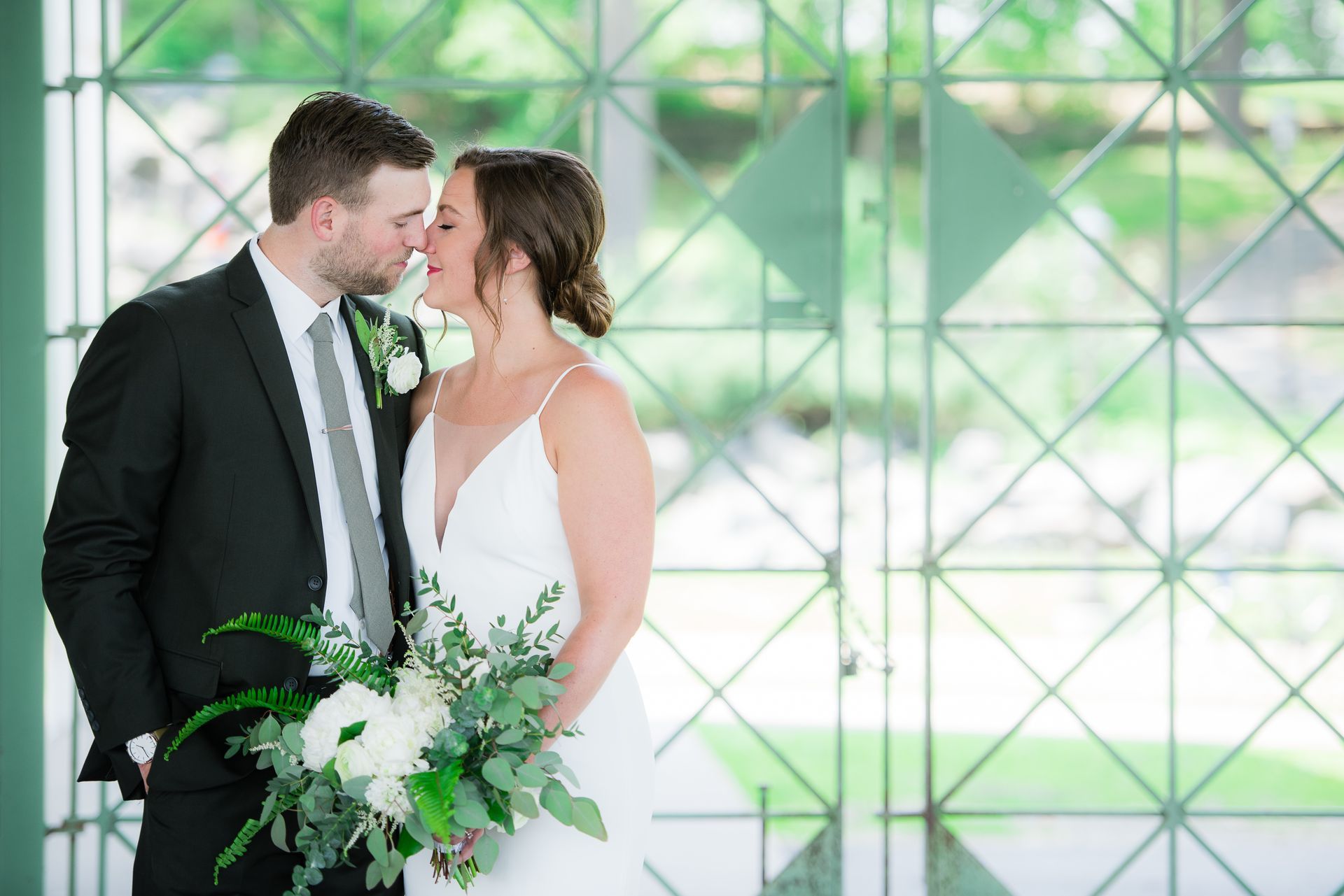 Couple embracing at a wedding; man in black suit, woman in white dress, holding bouquet, in front of a window.