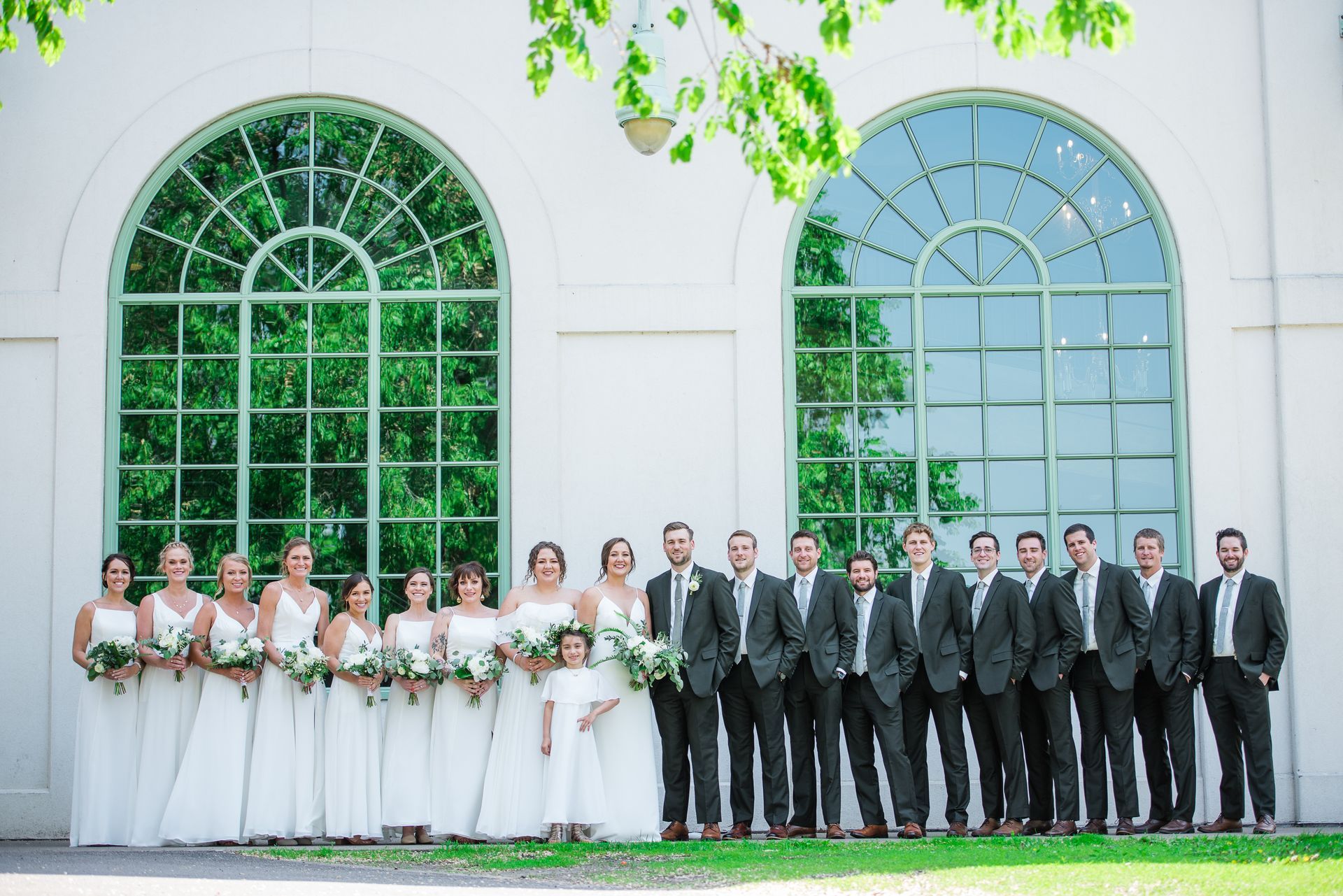 Wedding party poses in front of large arched windows with greenery; bridesmaids in white, groomsmen in black suits.