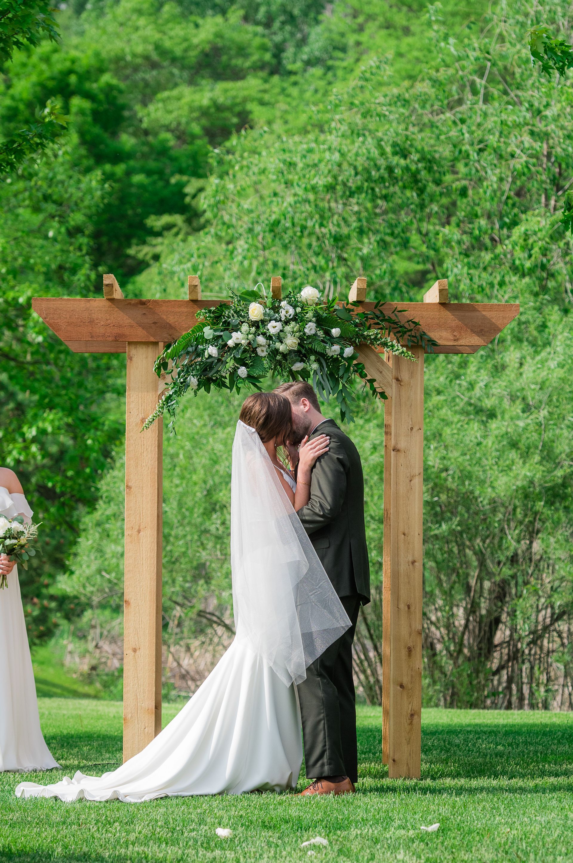 Bride and groom kissing under a wooden arbor decorated with flowers, outdoors in a green space.