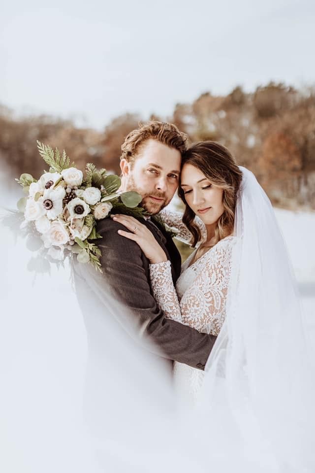 Bride and groom embrace in snow. He wears a dark suit, she a white lace dress and veil, holding bouquet.