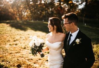 Bride and groom walking in a field, holding hands. Bride in white dress, groom in suit. Golden sunlight.