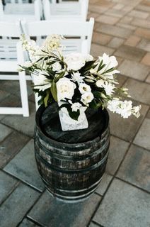 White floral arrangement on a wooden barrel; two white chairs and tiled floor in the background.
