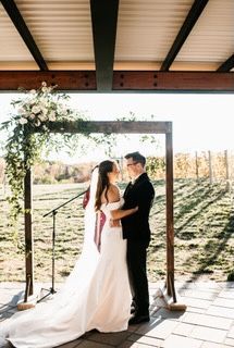 Bride and groom embrace under a floral arch during outdoor wedding ceremony. Vineyard in the background.