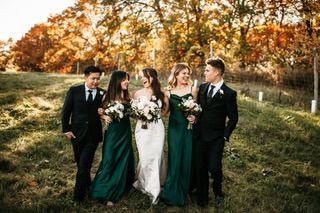 Wedding party walking through a field with fall foliage; bride in white gown, bridesmaids in green dresses, groomsmen in black suits.