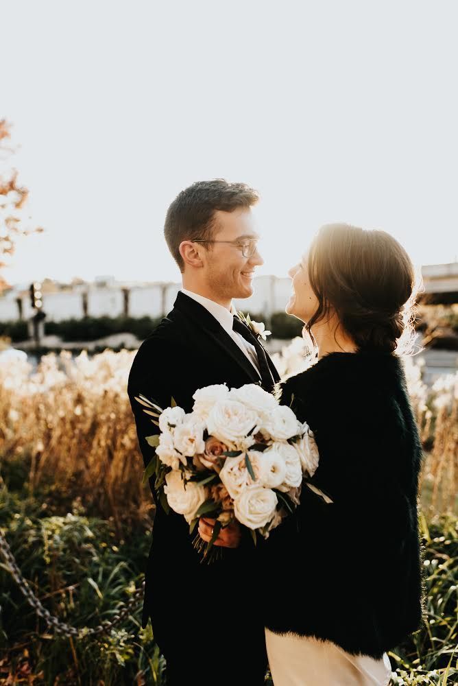 Wedding couple in embrace, woman holding flowers, smiling at the man outdoors with sunlit background.