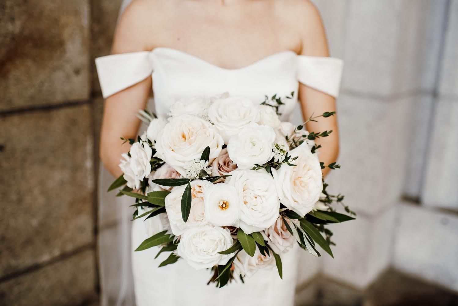Bride in white off-the-shoulder dress holding a bouquet of white and blush roses with greenery.
