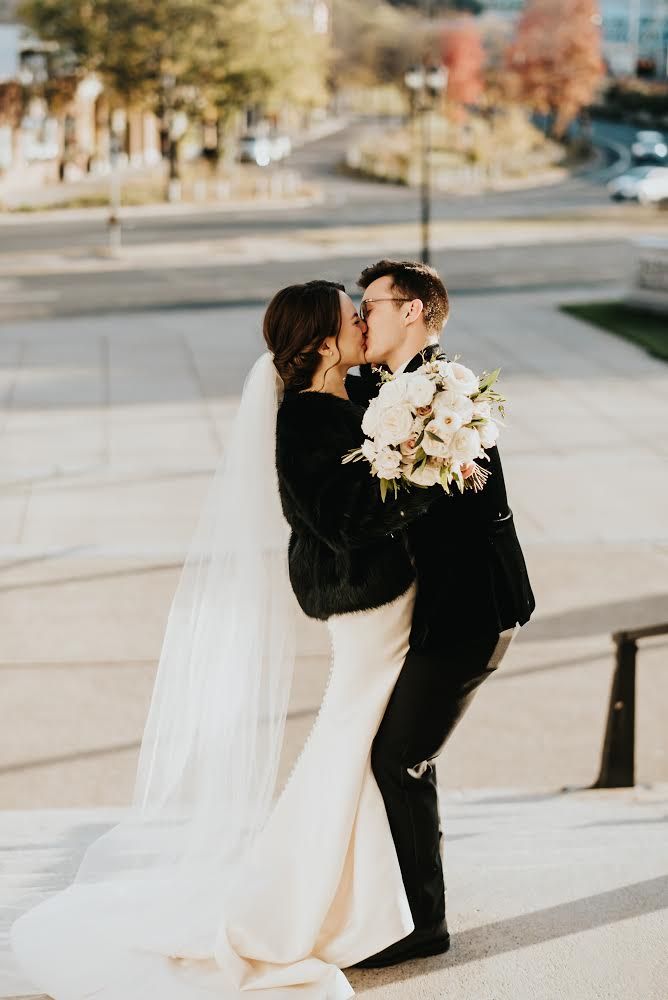 Bride and groom kiss on outdoor steps; bride in white gown and fur, groom in black suit.