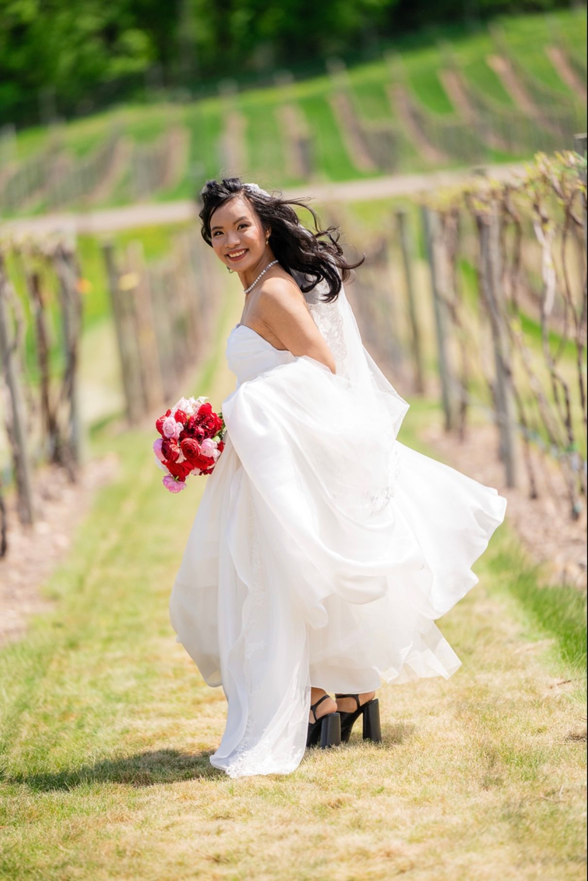 Bride in white dress holding bouquet, smiling, walking in vineyard.