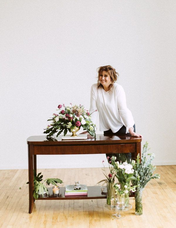 Bridesmaids in gray and blush dresses hold bouquets, standing behind the bride in a white gown on a red carpet.