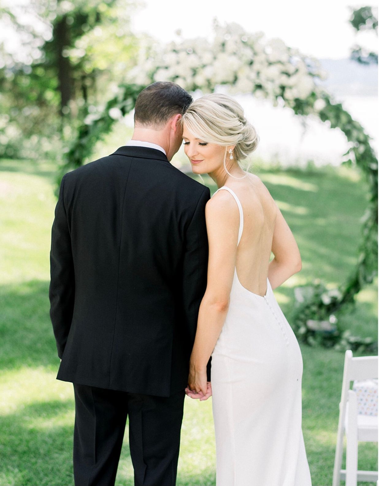 Bride and groom embrace at an outdoor wedding ceremony. Bride in white dress, groom in black suit, floral arch.