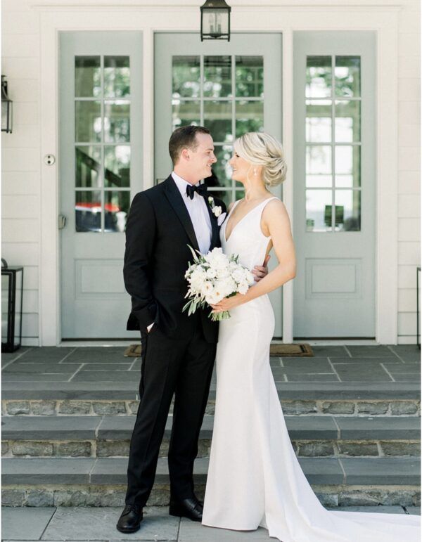 Wedding couple: man in black tux and woman in white gown, embrace on steps in front of a house.