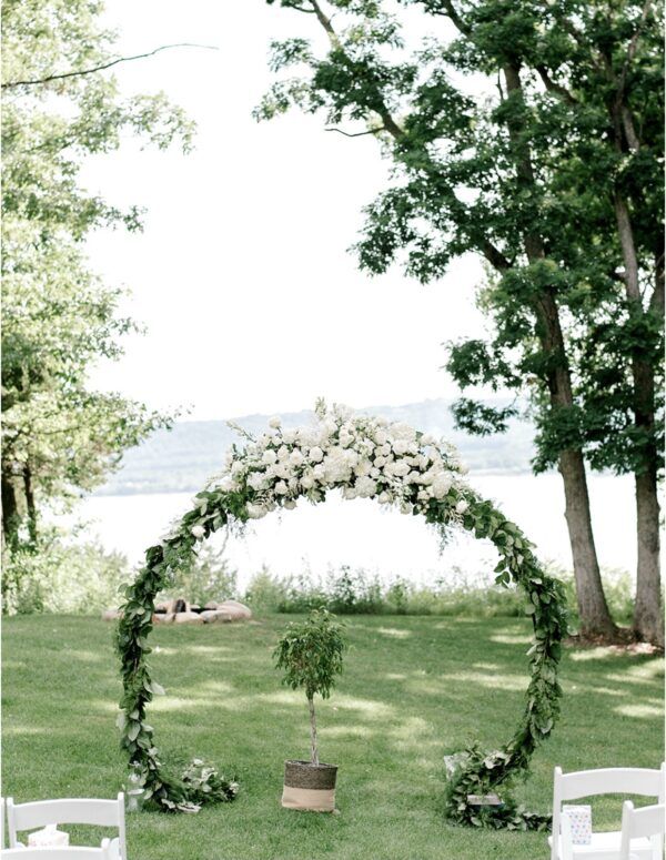 Wedding arch with white flowers and greenery, set on a green lawn with trees and a lake in the background.