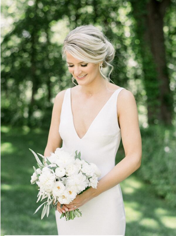 Bride with updo hairstyle, smiling, holding white floral bouquet, wearing white dress in a garden setting.