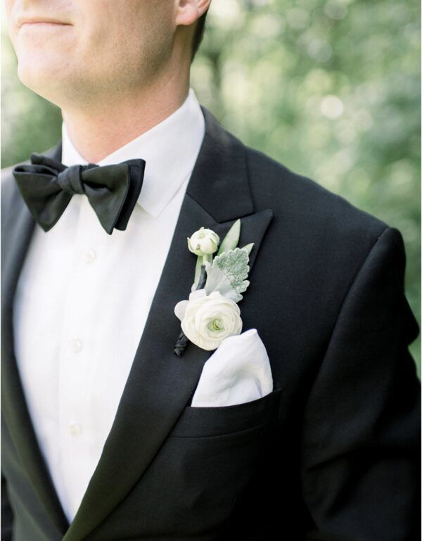 Man in black tuxedo, white shirt, bow tie, pocket square, boutonniere, light background.