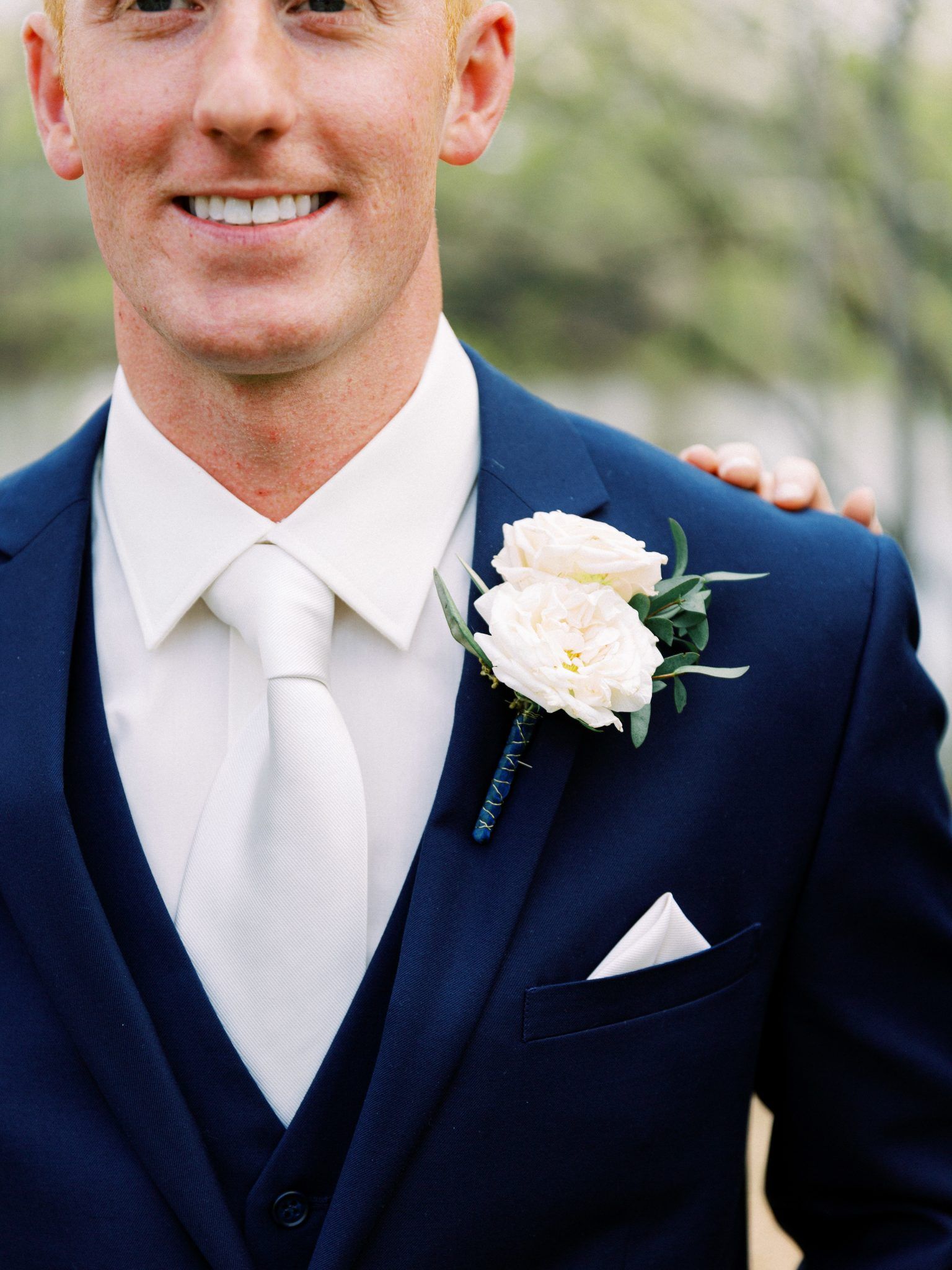 Man in navy suit with white tie and boutonniere smiles. Outdoors setting.