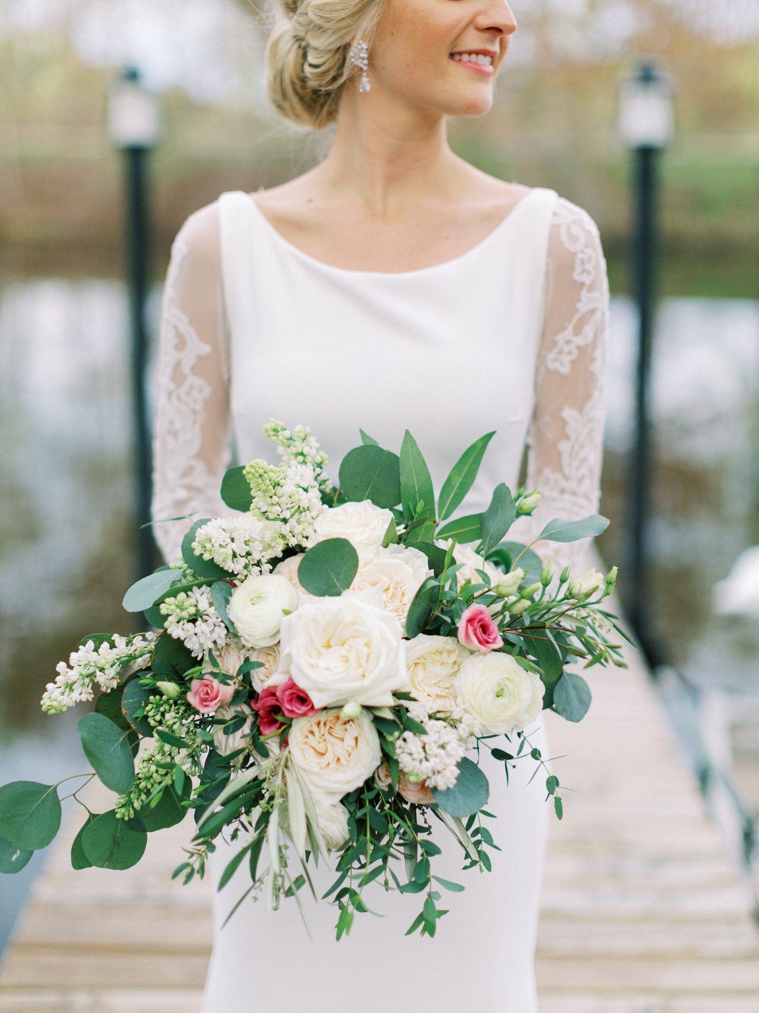 Bride in white gown holding bouquet of white and pink flowers, standing on a wooden dock.