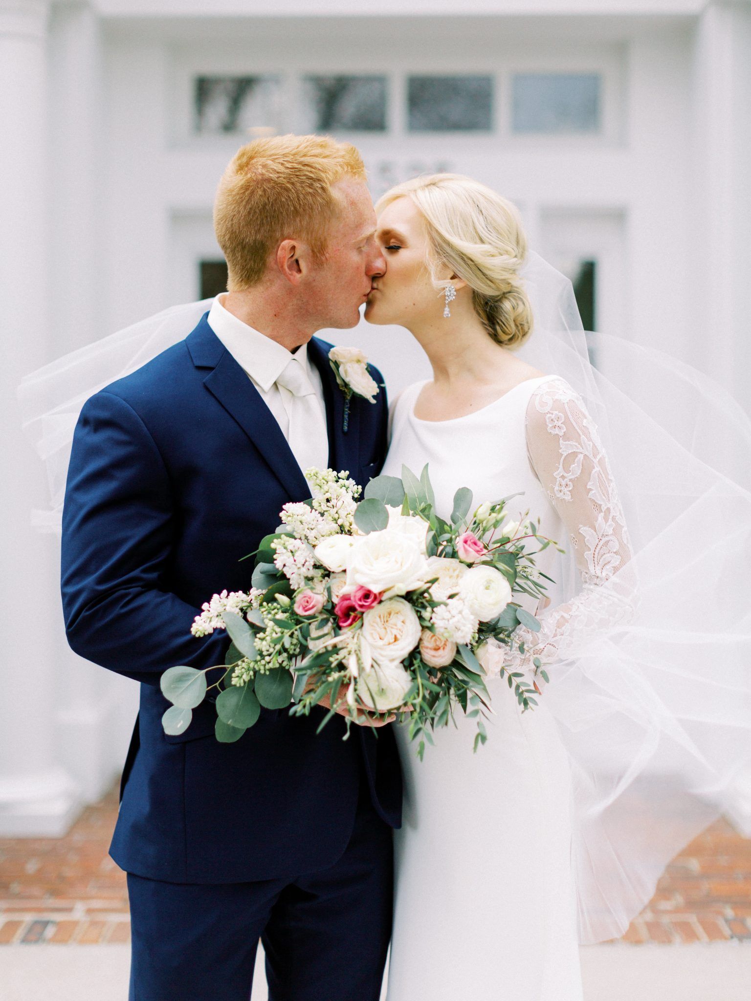 Wedding couple kissing; man in blue suit, woman in white dress, holding bouquet.