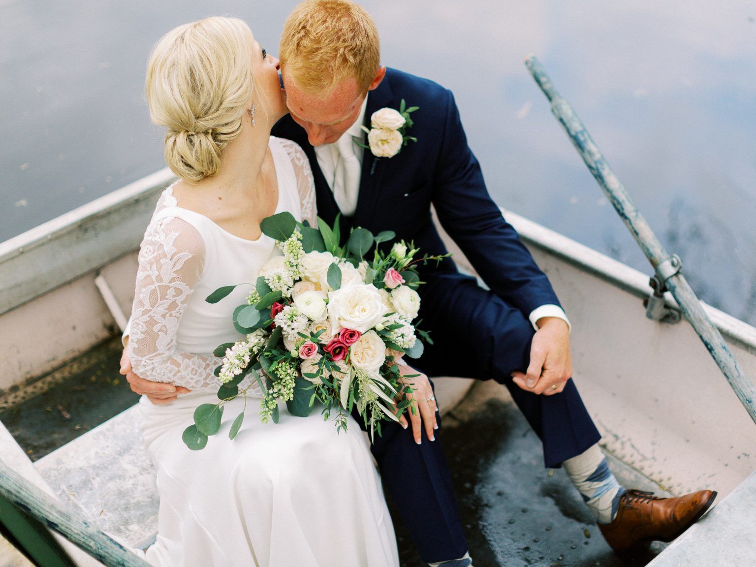 Bride kissing groom in a boat on the water; both are dressed in wedding attire.