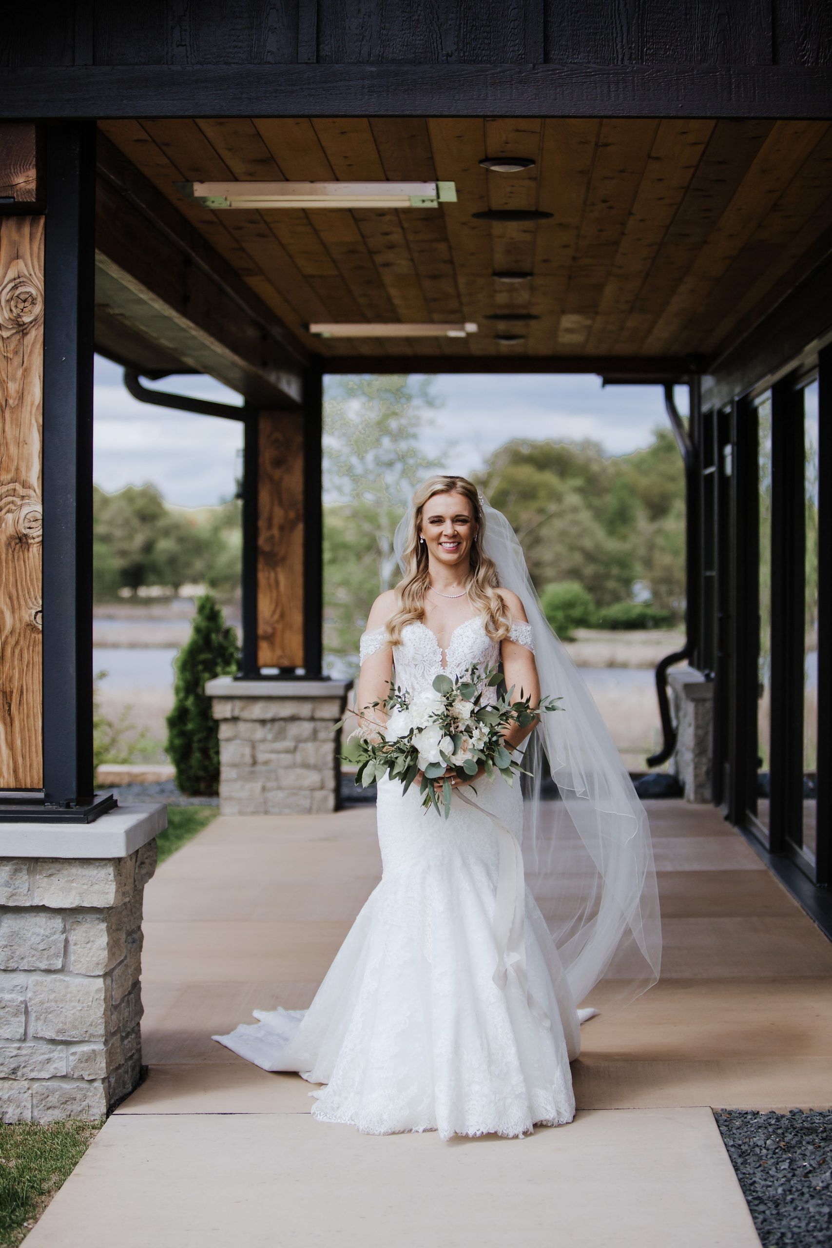 Bride in white dress holding bouquet, smiling, standing under wooden canopy.