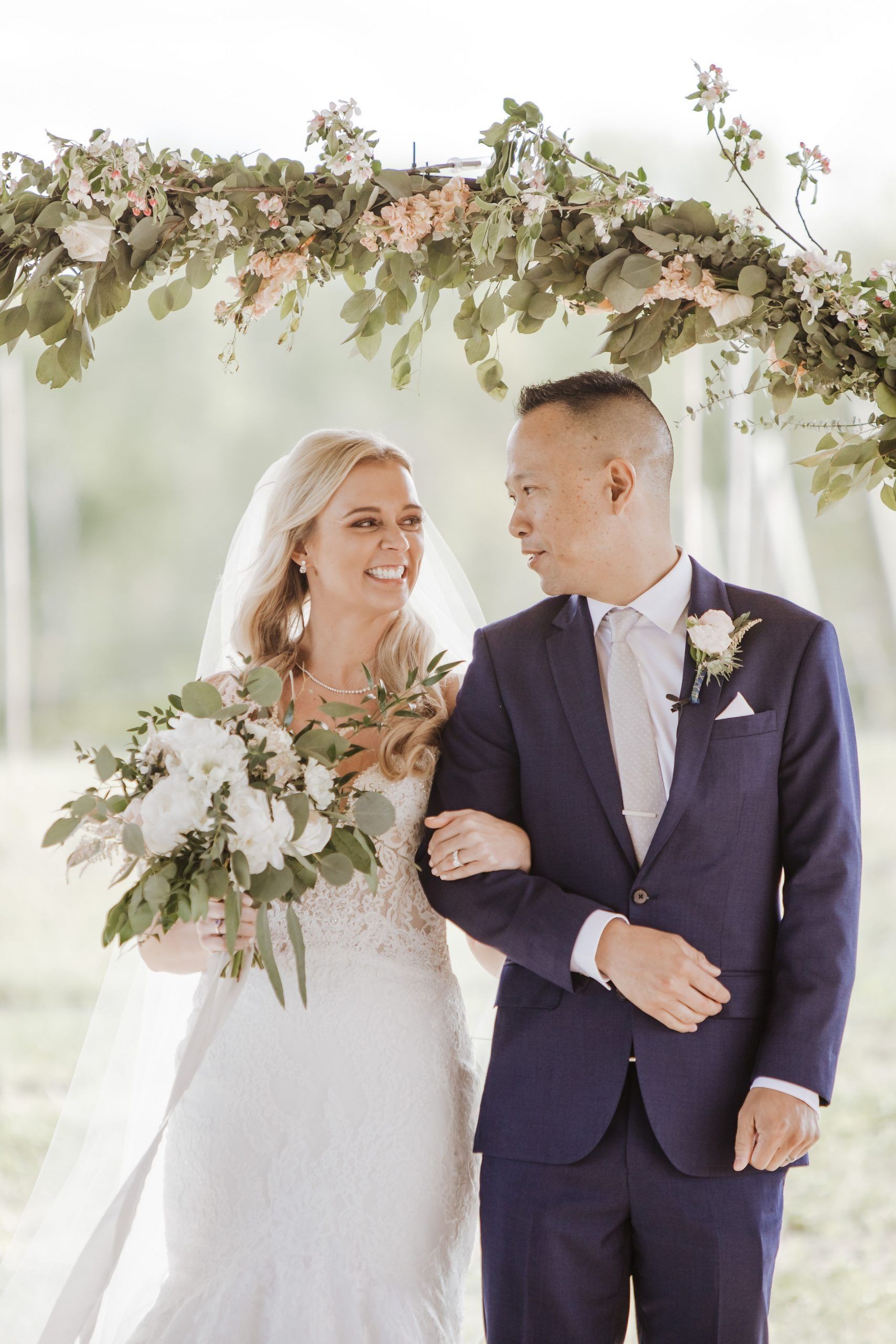 Bride and groom arm-in-arm under a floral arch after wedding. Smiling; woman holds bouquet.