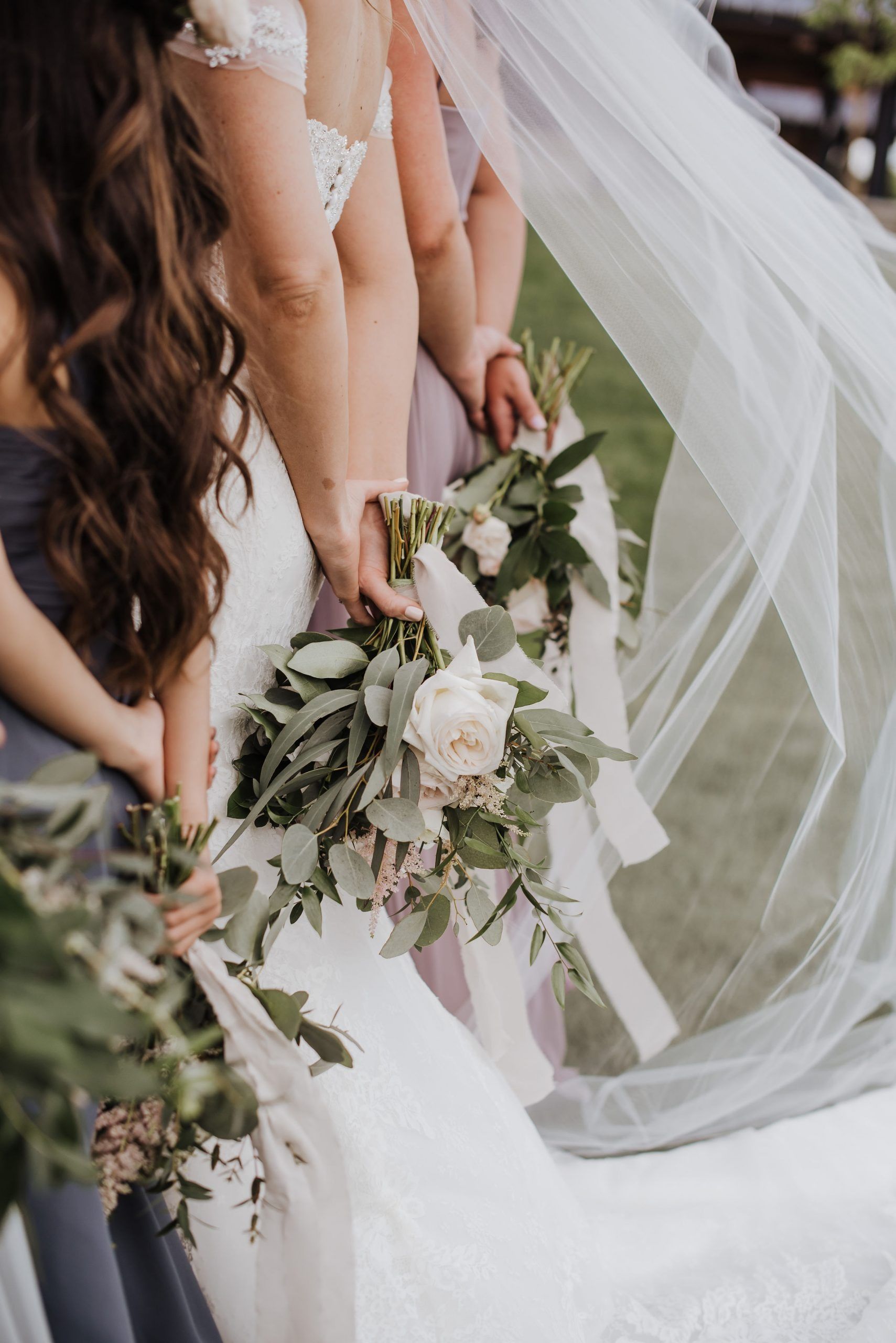 Bridesmaids holding bouquets of flowers, one wearing a white wedding dress, outside in a grassy area.