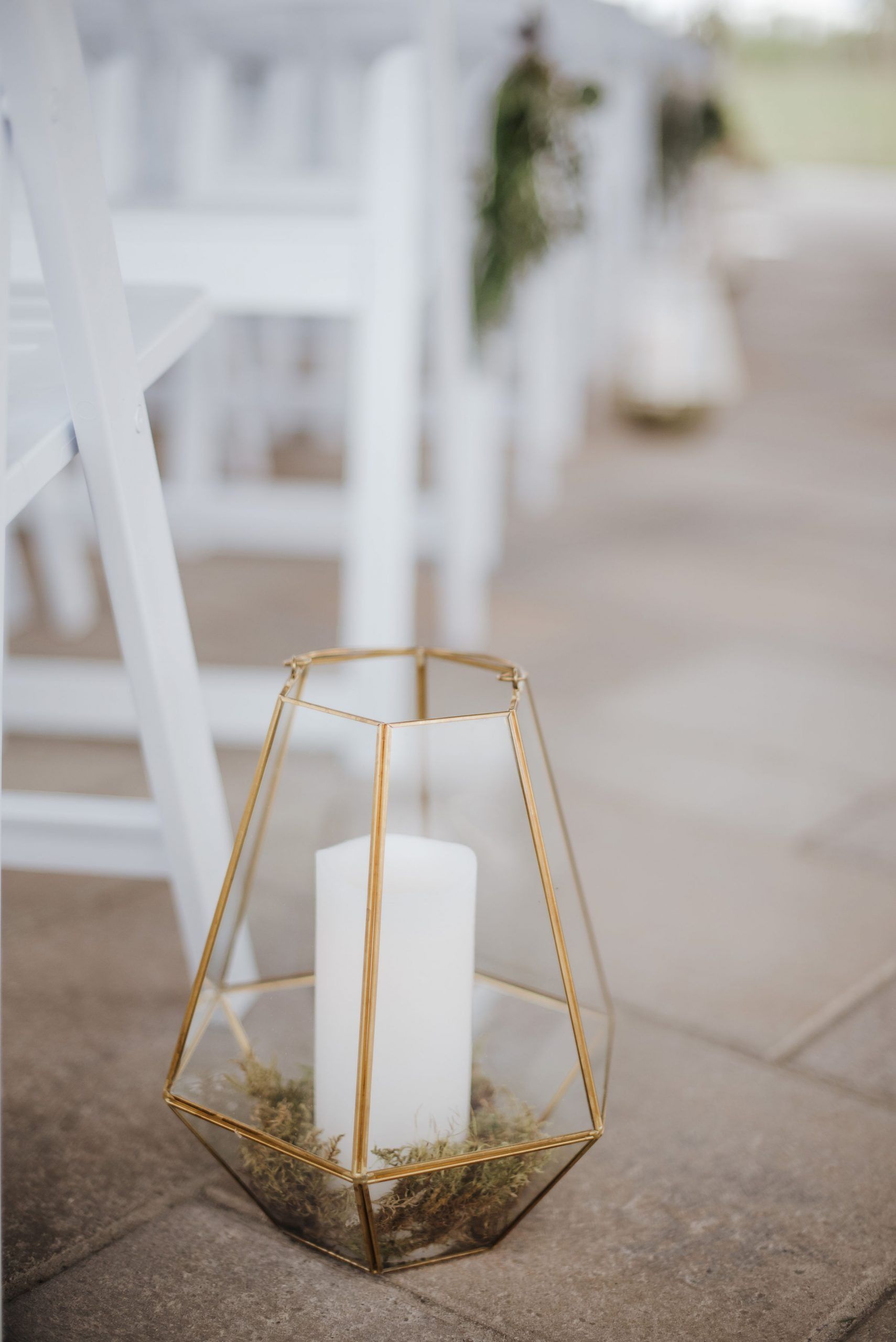 Gold geometric lantern with white candle, on stone path, near white chairs.