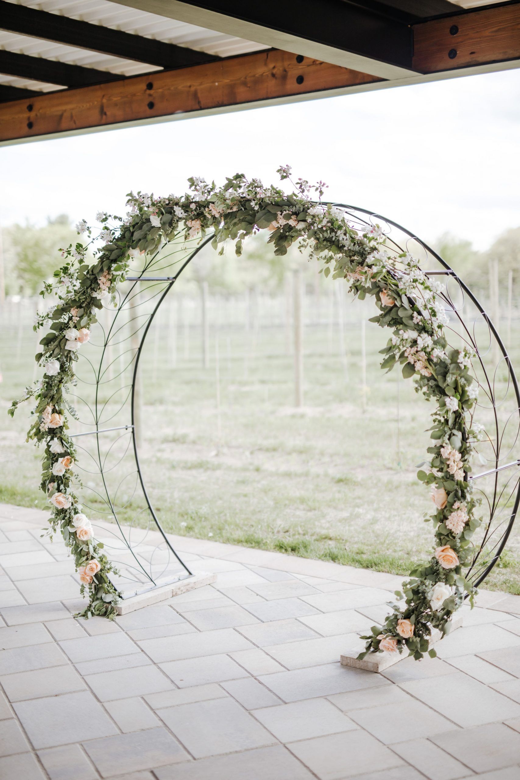 Floral wedding arch with peach and green blooms set on a brick patio, vineyard backdrop.