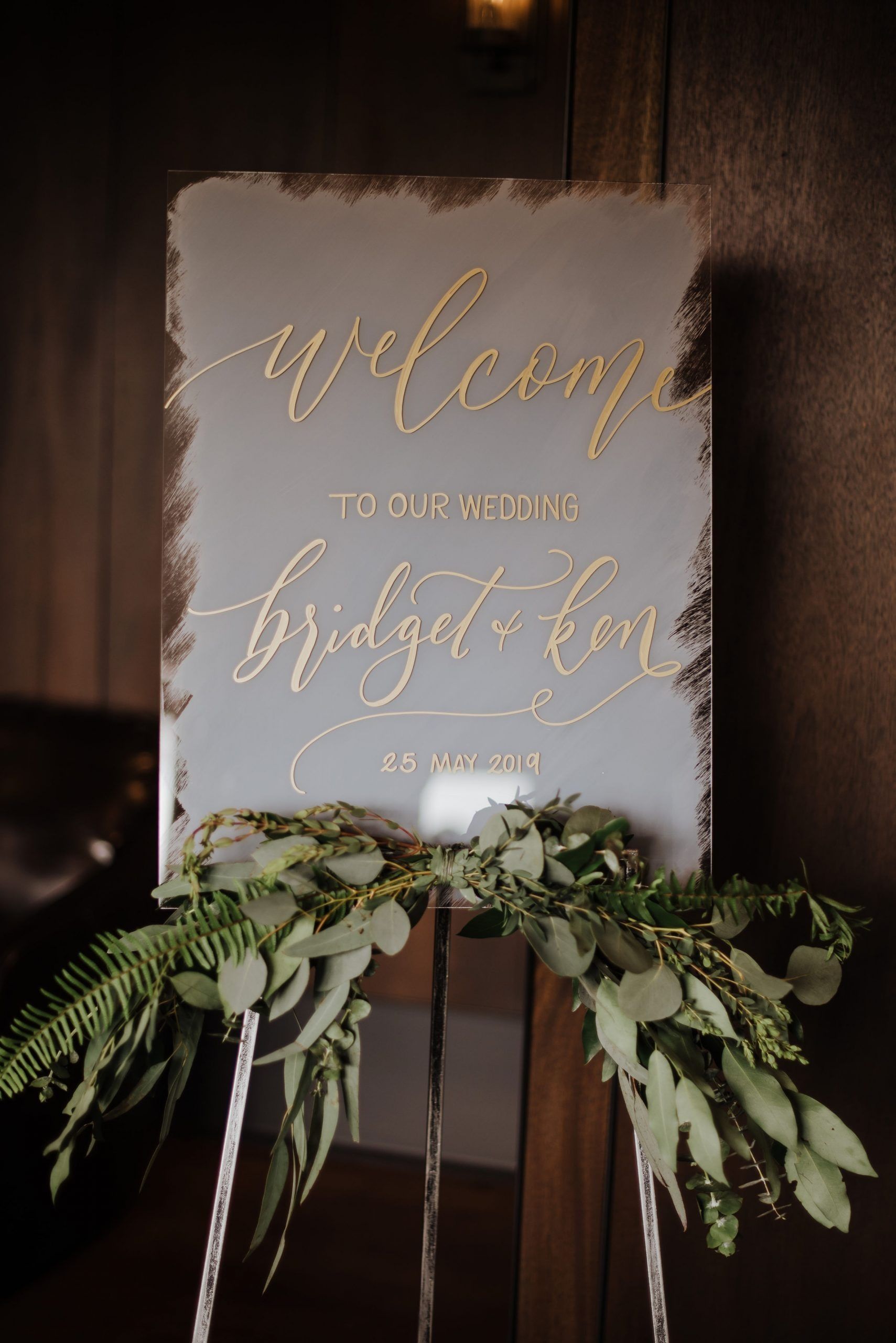 Welcome sign with gold lettering on a grey background, decorated with greenery, on a wooden easel.