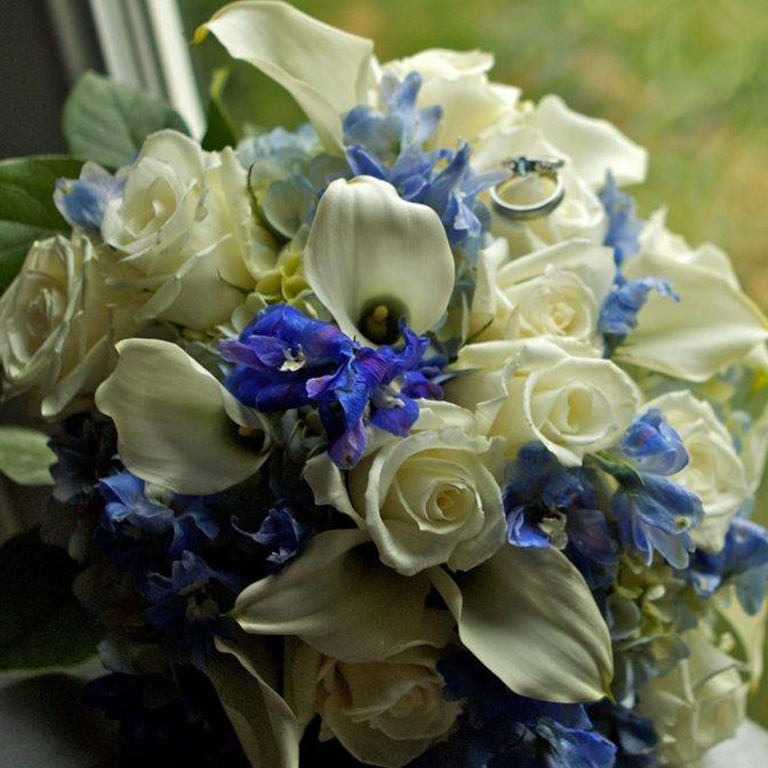 Wedding bouquet with white roses, blue flowers, and calla lilies. A wedding ring rests on top.