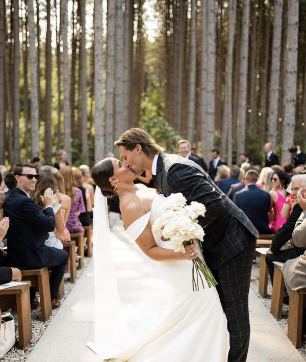 Newlyweds kissing in a forest ceremony, the bride in white, groom in a suit, guests seated.