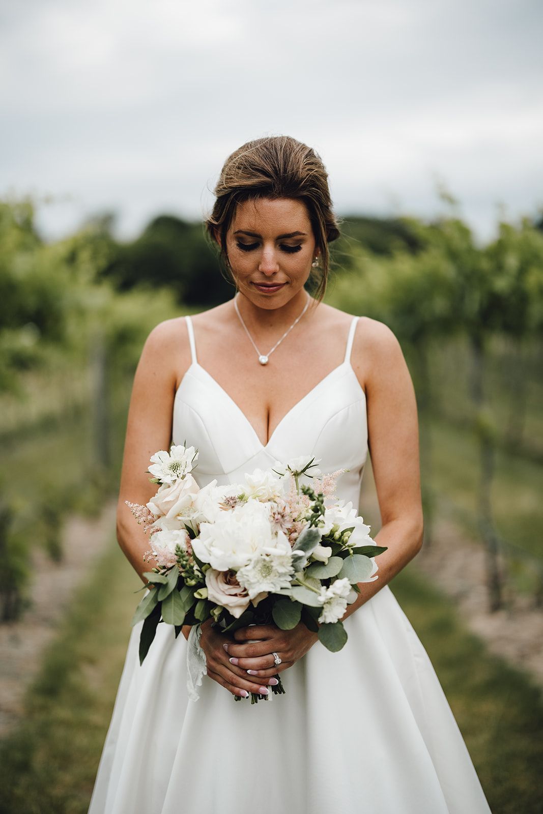 Bride in white gown holding bouquet, looking down; vineyard background.