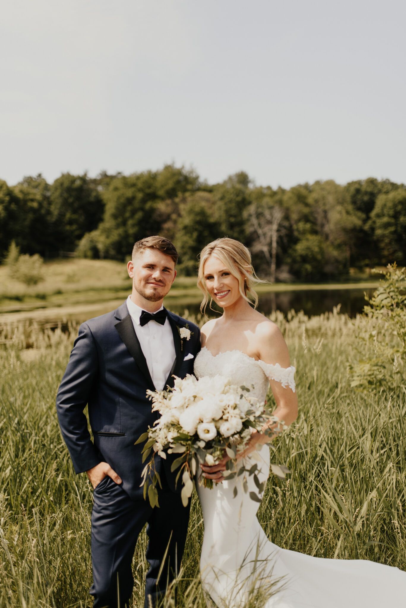 Wedding couple poses in a field. The groom wears a blue suit, and the bride wears a white off-the-shoulder dress.