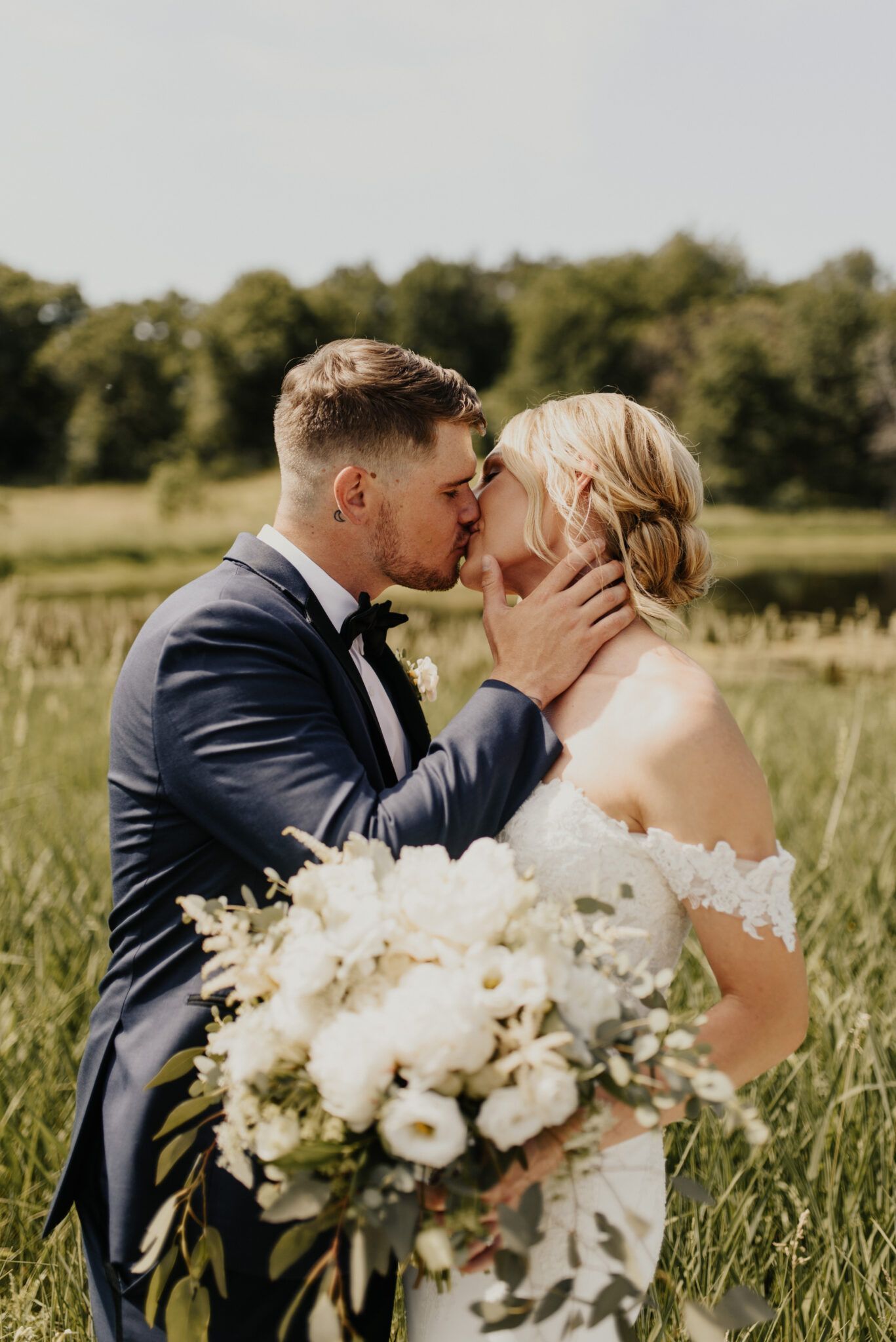 Bride and groom kissing in a field, bride holding bouquet. Groom in blue suit, bride in white dress.