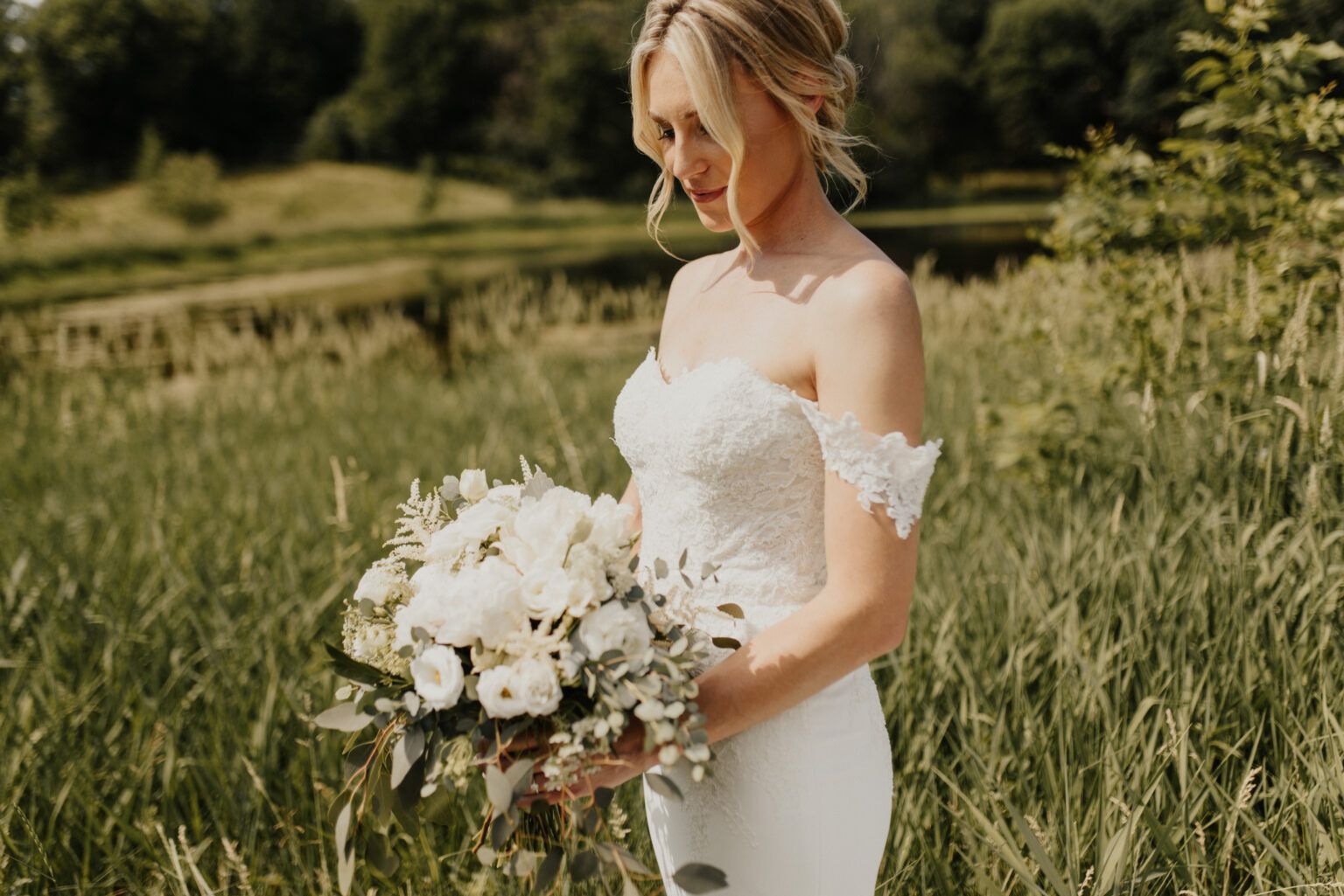 Bride in strapless white gown, holding bouquet, standing in grassy field, gazing down.