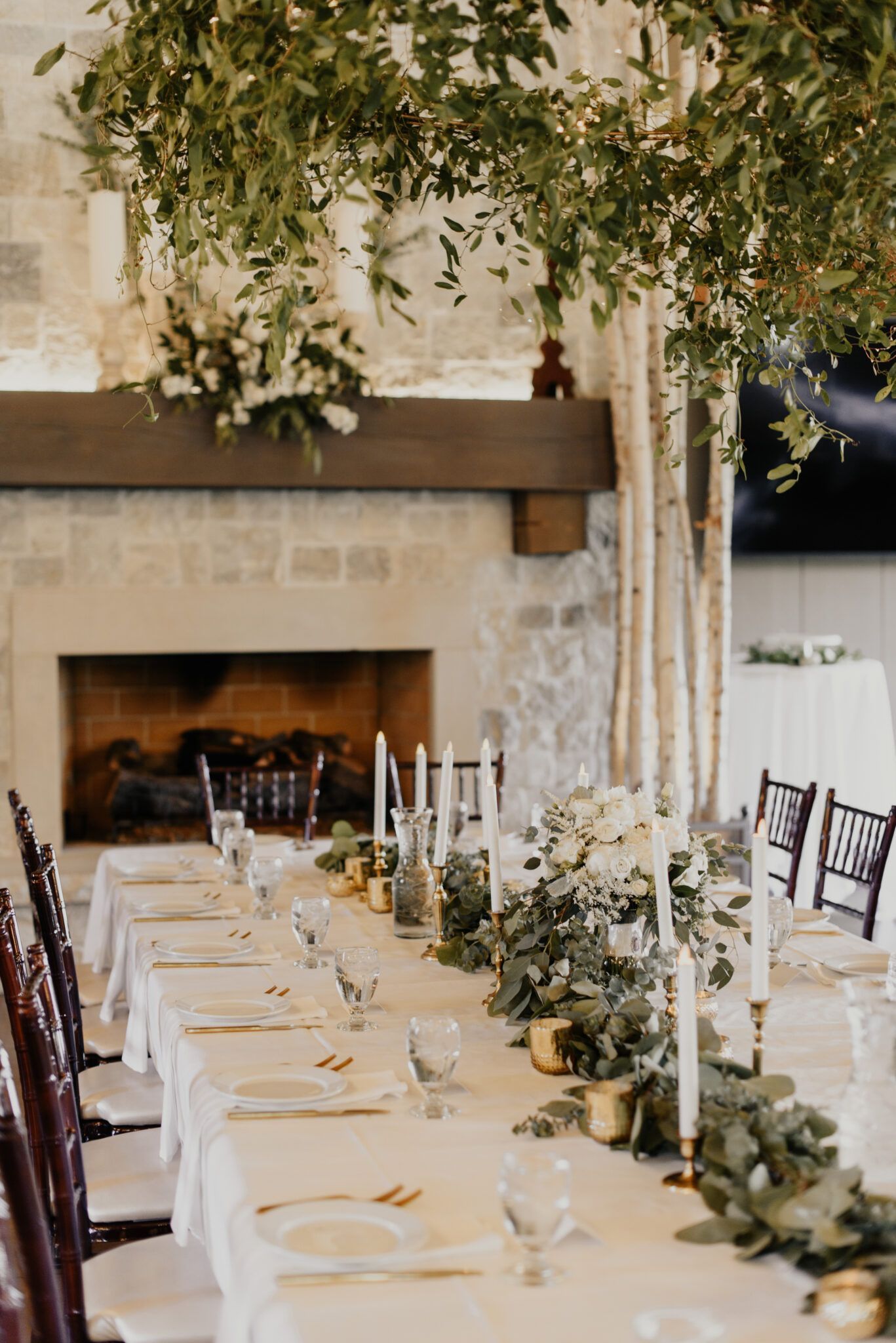 Formal dining table set for a wedding with white linen, gold accents, and greenery. Fireplace in background.