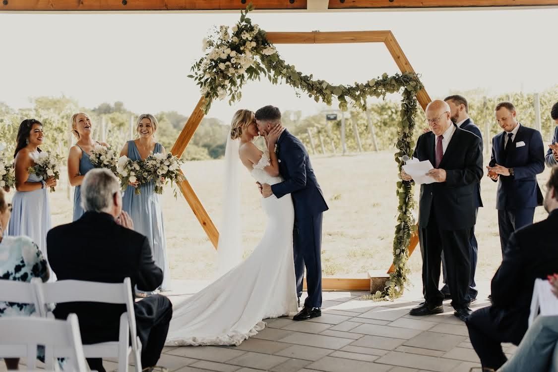 Bride and groom kiss under floral arch at outdoor wedding ceremony.