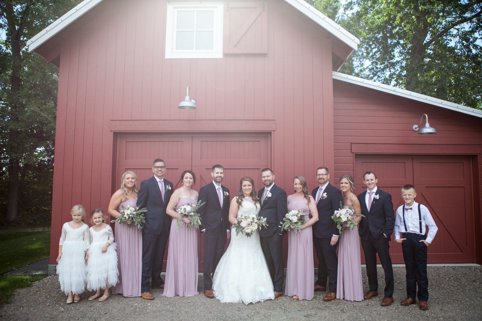 Wedding party poses in front of a red barn; bride in white dress, bridesmaids in lavender, groomsmen in navy.