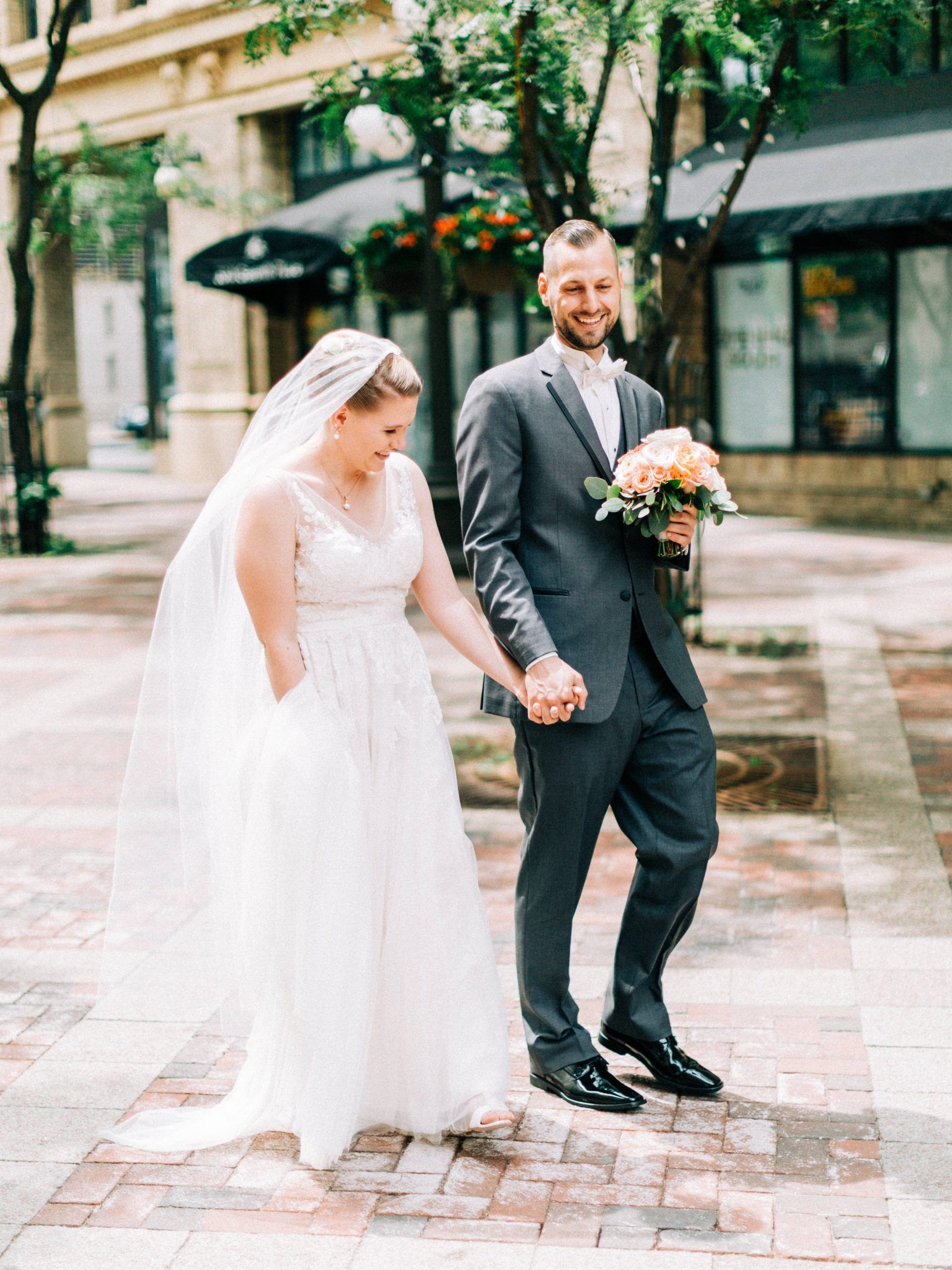 Bride and groom walking hand-in-hand on a brick street; she's in a white dress, he's in a gray suit.
