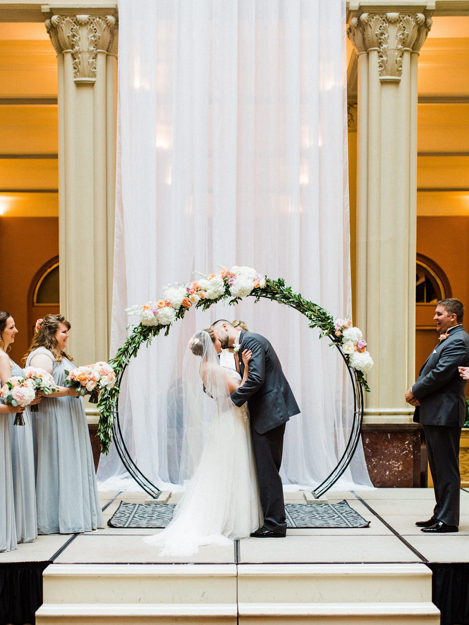 Bride and groom kissing under a floral archway during a wedding ceremony, with bridesmaids and a groomsman in the background.